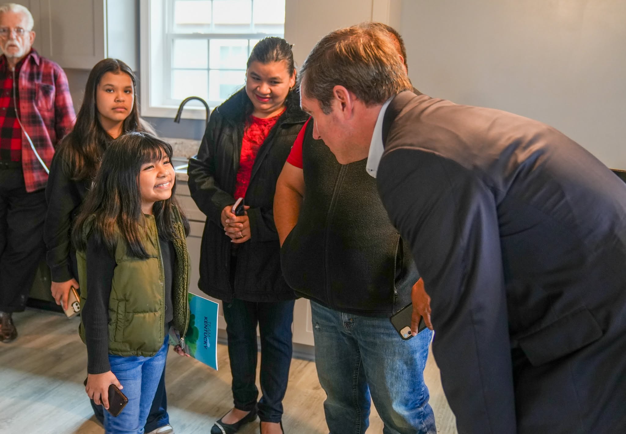 Governor leans down to say hello to young girl with long brown hair smiling at him in her new home on high ground.